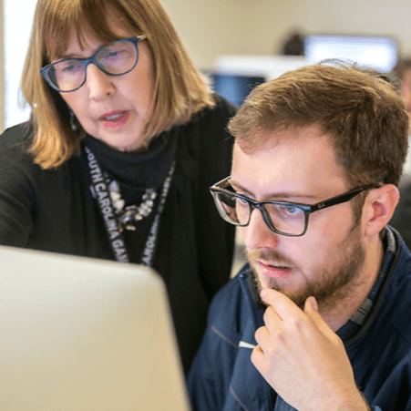 Male student working at computer with teacher looking over shoulder to help