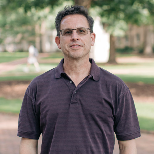 Man with glasses and short brown hair smiling, wearing a dark brown polo t-shirt