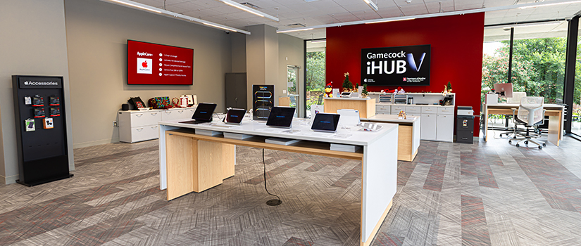 Interior of the Gamecock iHub featuring a central table with laptops on display, a red accent wall with the Gamecock iHub logo, and large windows overlooking an outdoor seating area.