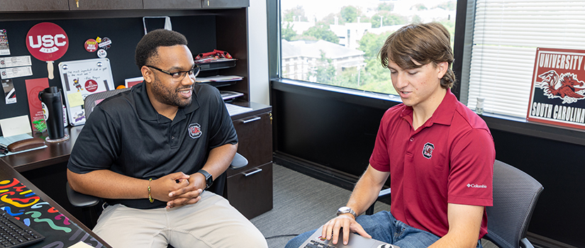 An advisor sits with a student in his office while the student looks at a laptop