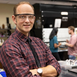 Morgan Stefik poses in his lab, where two graduate students discuss their battery research in the background